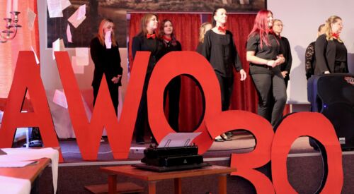 Bühne. Frauen in schwarzer Kleidung und roten Krawatten Fliegen. Auf der Bühne große rote Buchstaben und Ziffern: AWO 30